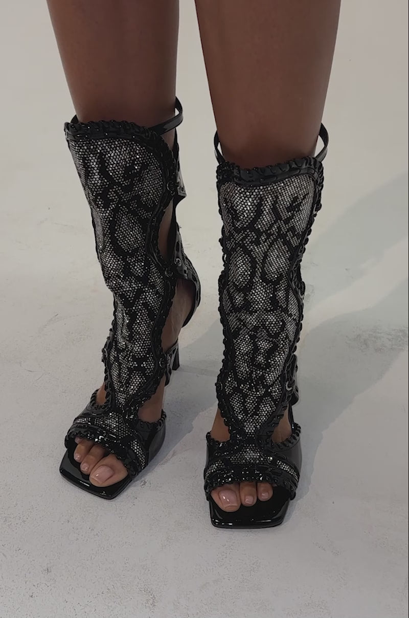 Video of woman modeling the AZALEA WANG SOLANGE SILVER SNAKE RHINESTONE GLADIATOR SANDAL, featuring black, textured material with lace-up stiletto heels, cutout details, and intricate design elements.