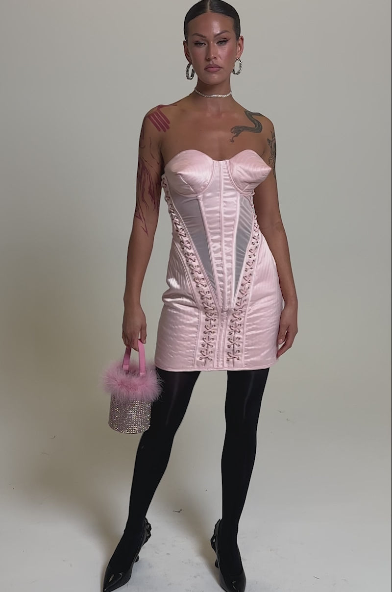 A video of a woman modelling the WOAH BABY SATIN MINI DRESS WITH CONE DETAIL, paired with black sheer tights, black heels, and a pink fluffy handbag. 