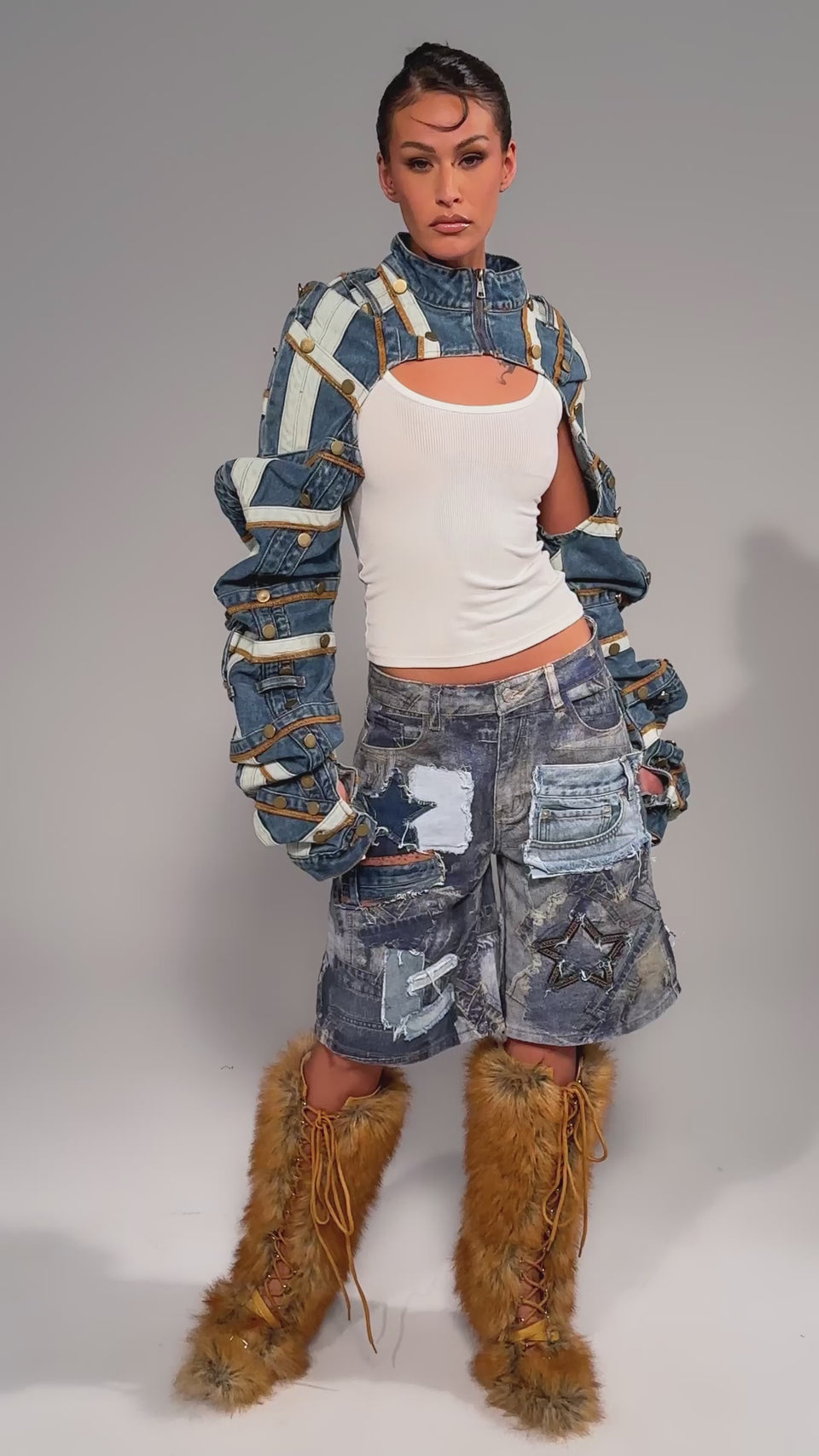Video of woman modeling the STACKED UP DENIM BOLERO with cut-out sleeves over a white tank top and distressed patchwork jean shorts, standing against a plain gray background.