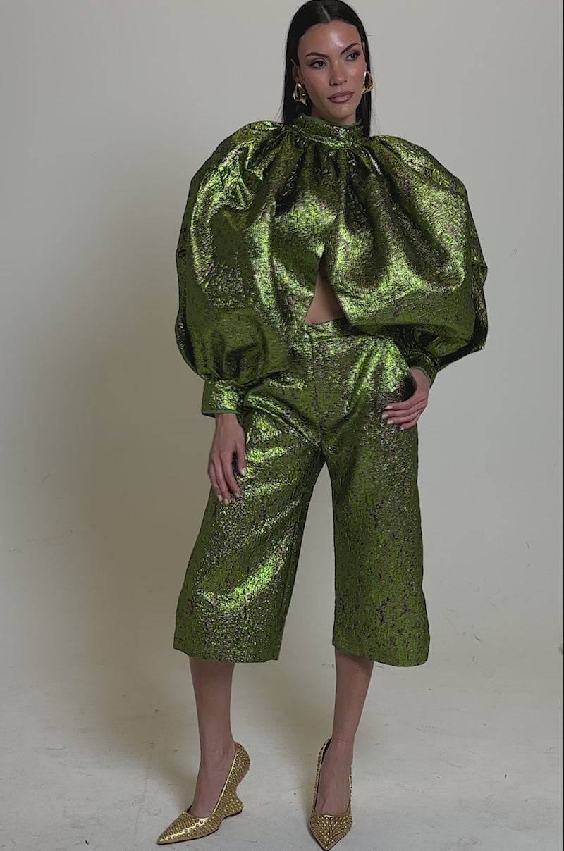 A video of a woman modelling the LAYLA BROCADE PUFF SLEEVE MOCK NECK TOP in shiny green, paired with matching wide-leg cropped pants and gold pointed heels, standing out against a neutral background.