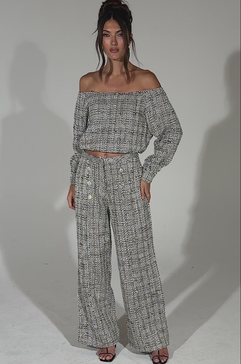 A video of a woman walking forward in black heeled sandals wearing the DRESSY VIBES TWEED PANT, a high-rise, wide-leg silhouette paired with an off-the-shoulder, long-sleeve patterned top against a plain light background.