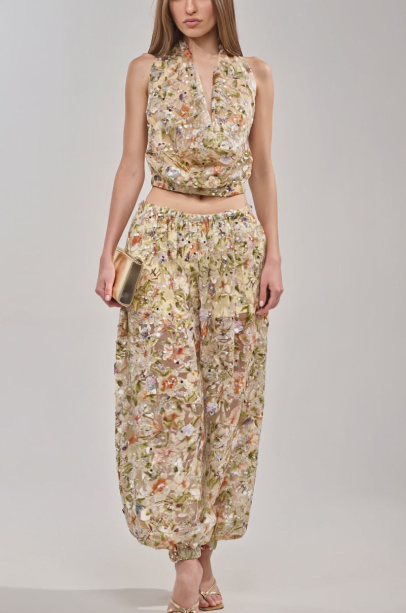 A video of a woman wearing the LETS TALK ABOUT IT BALLOON PANT, a floral-patterned, gathered-ankle pant, with a matching sleeveless halter top, gold high-heeled sandals, and a small beige clutch.