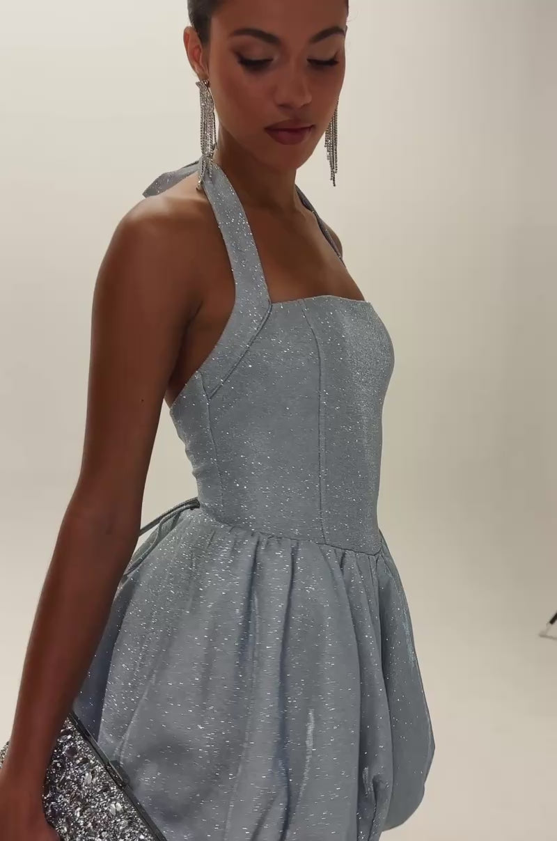 Video of woman modeling the GLISTEN UP SHIMMER HALTER MINI DRESS, a light blue sparkly mini dress with a bubble skirt. She holds a silver clutch, wears long earrings, and has sleek hair with a confident look.