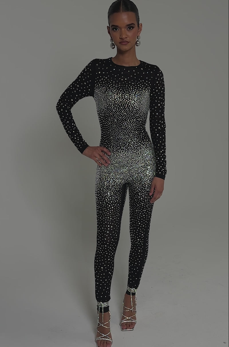 Video of woman modeling the ALL ABOUT ME RHINESTONE JUMPSUIT, a sparkling, curve-hugging outfit paired with silver earrings and high-heeled sandals against a gray background.