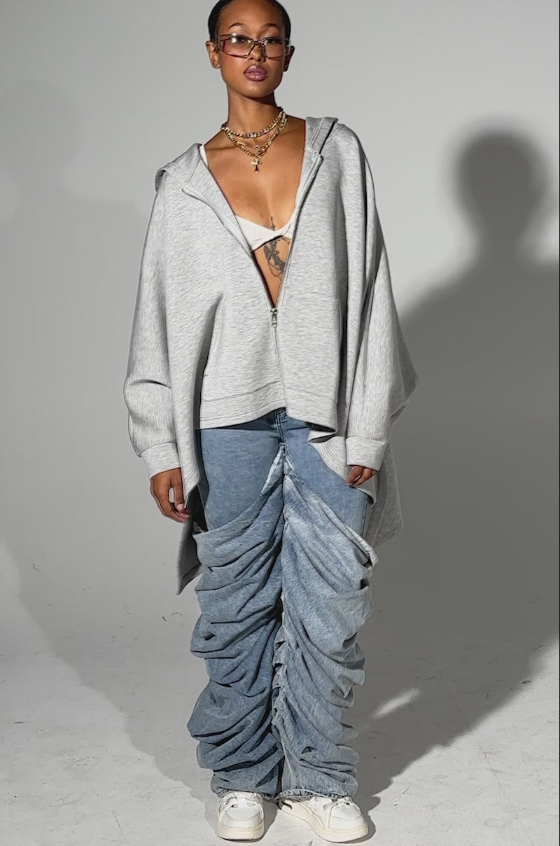 A video of a person wearing the CREW LOVER OVERSIZED ZIP UP SWEATSHIRT in heather grey over a white bralette, styled with blue ruched baggy jeans and white sneakers. They accessorize with glasses and layered necklaces against a plain light background.