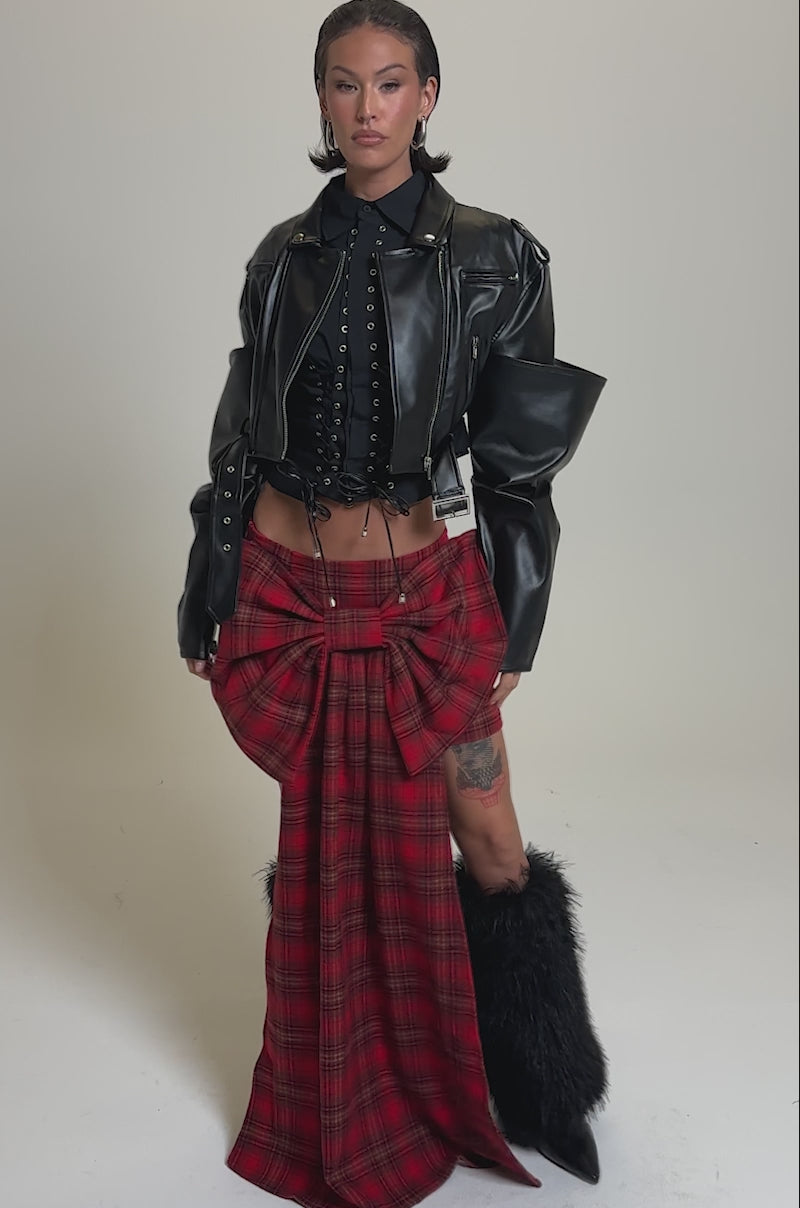 A video of a model wearing the STACK IT UP CROPPED MOTO jacket in black faux leather, styled with a lace-up top, red tartan skirt with a large bow and panel, black furry boots, and visible leg tattoos against a plain background.