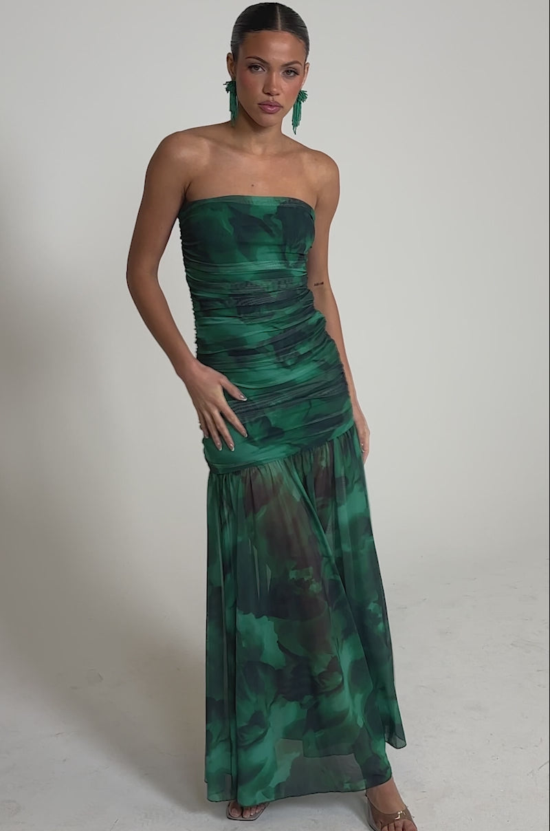 Video of woman modeling the UNDER THE SEA MAXI DRESS—a strapless mesh gown with a ruched bodice, green and black patterns, and a sheer flowing skirt. Her hair is pulled back, she accessorizes with green earrings and neutral heels against a light backdrop.