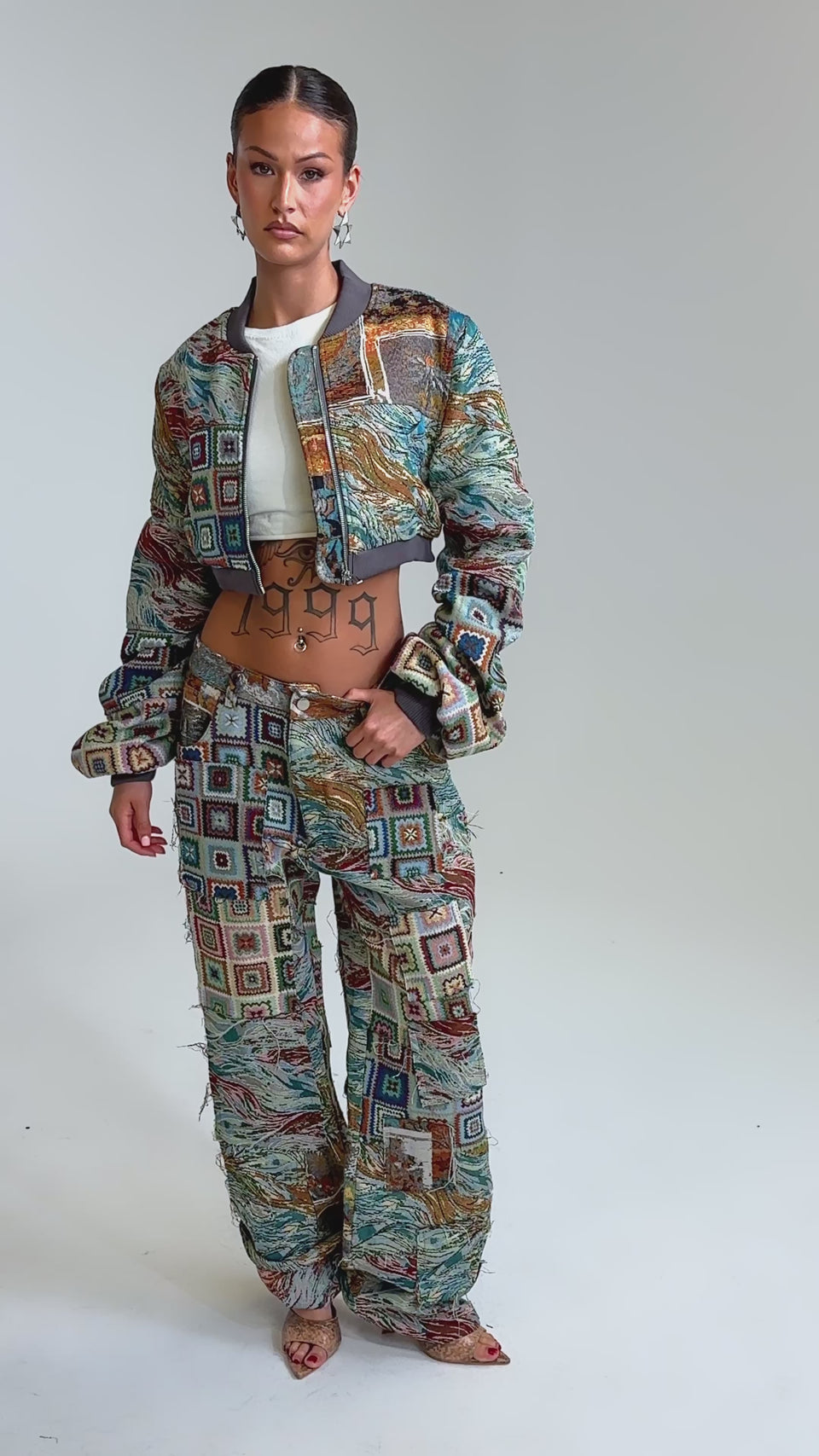 Video of woman modeling the WILD CROPPED PATCHWORK BOMBER and matching pants, featuring bold square and wave patterns. A "1999" tattoo, hoop earrings, and slicked-back hair complement the jacket’s vibrant tapestry texture.