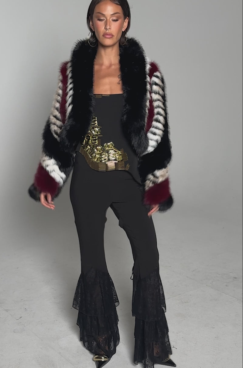A video of a person in the HARD TO FIND PANELED FAUX FUR jacket with bold black, white, and red patterns, paired with a black strapless top, flared lace pants, gold belt, and black pointed shoes against a plain gray background.