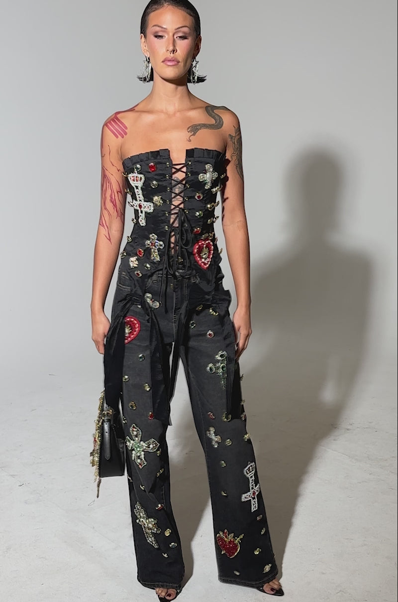 A video of a person wearing the MAEVE EMBELLISHED CORSET—strapless and decorated with colorful jeweled patches and cross motifs—paired with matching pants, and holds a black handbag featuring gold chain accents.