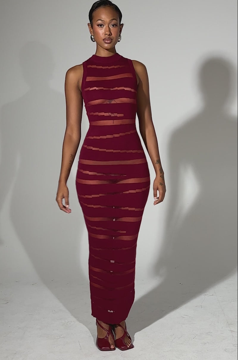 A video of a woman with short hair and minimal jewelry posing against a plain background in the CUT IT OUT SLEEVELESS KNIT MAXI DRESS IN BURGUNDY, featuring sheer horizontal stripes, cut-out paneling, and paired with open-toe heels.