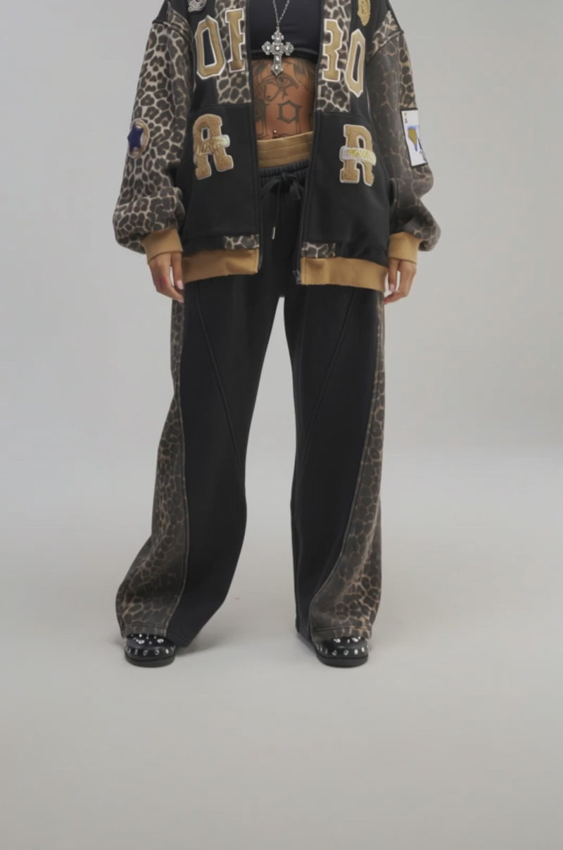 Video of a woman modeling the WORLD HOLD ON ZIP UP SWEATSHIRT IN MUSTARD—a cozy fleece varsity hoodie featuring a black and brown leopard print, gold lettering, heart patches, and other embroidered designs.