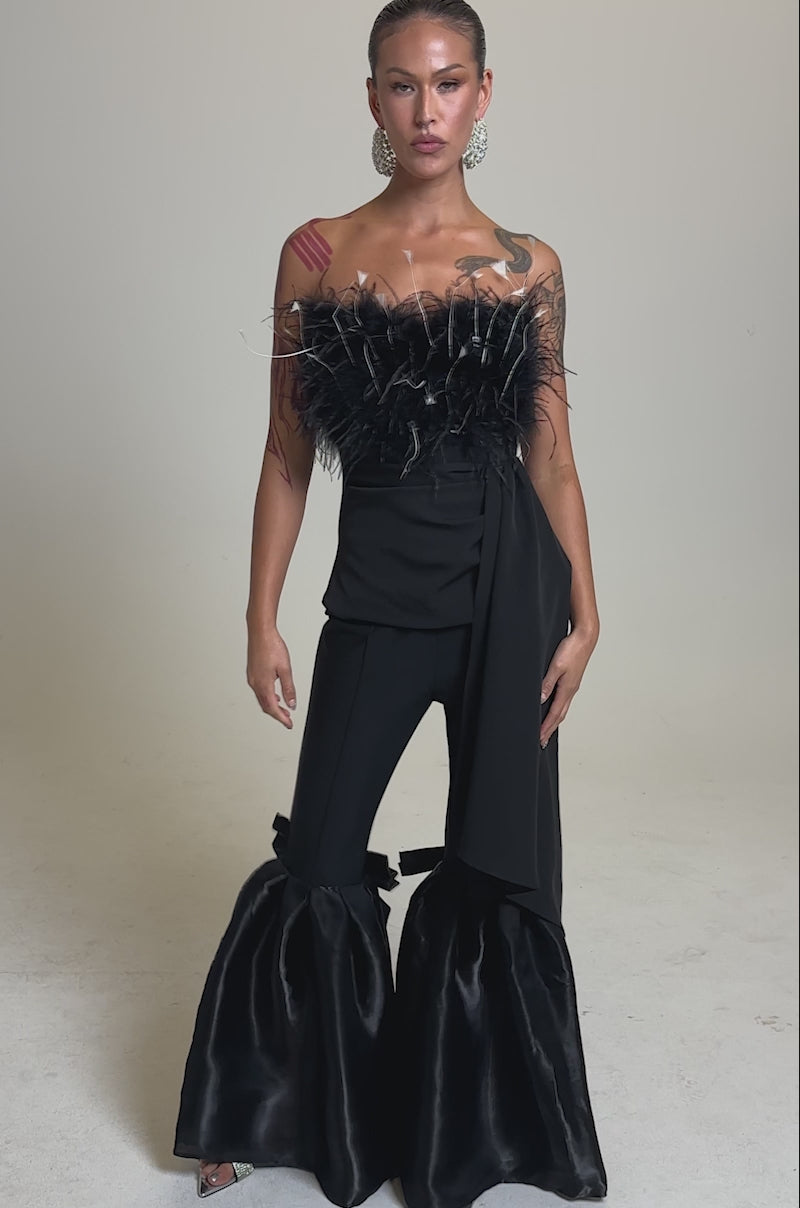 A video of a woman posing confidently in the CLARA FEATHER CORSET, a dramatic black strapless piece, paired with flared pants featuring large ankle bows and bold earrings, against a plain background.