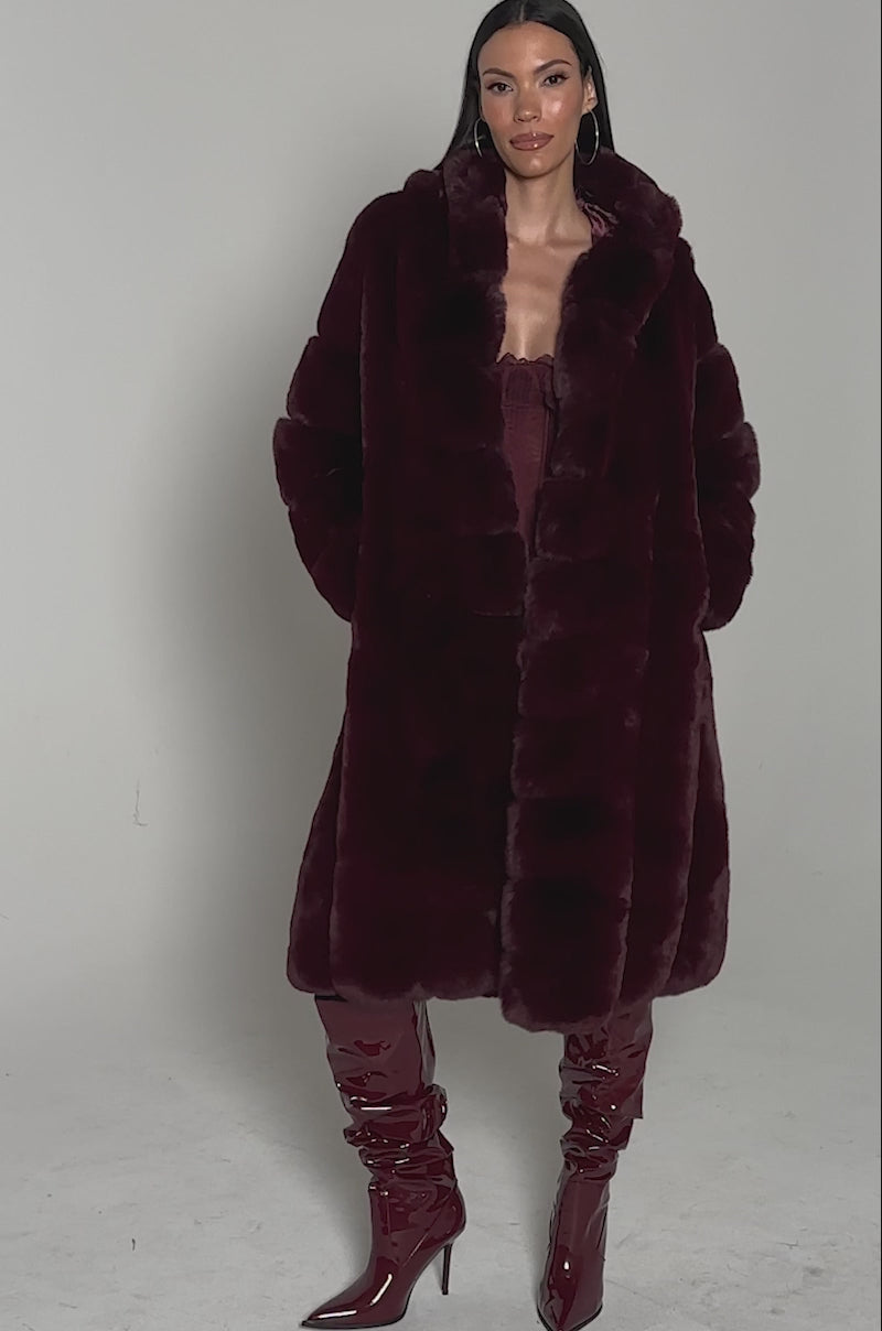 Video of woman modeling the BERLIN HOODED FAUX FUR IN BURGUNDY, styled with dark pants, matching glossy knee-high boots, hoop earrings, and sleek straight hair, standing against a plain light background.