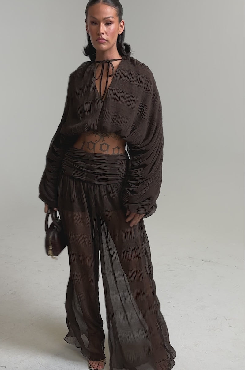 A video of a woman in the BABY THATS HOW I WANT IT BLOUSE, a sheer brown balloon top with a billowy cropped fit and matching wide-leg pants.