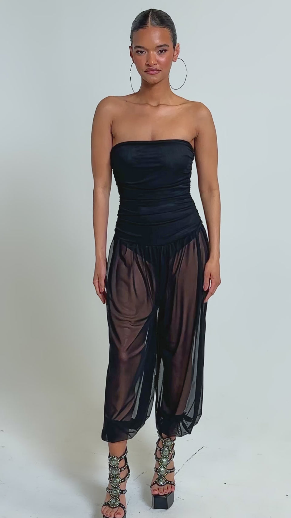 Video of woman modeling the NO CHILL MESH JUMPSUIT IN BLACK, featuring a strapless mesh bodice and sheer black harem pants with ruched ankles, accessorized with large hoop earrings, braided hair, and platform sandals.