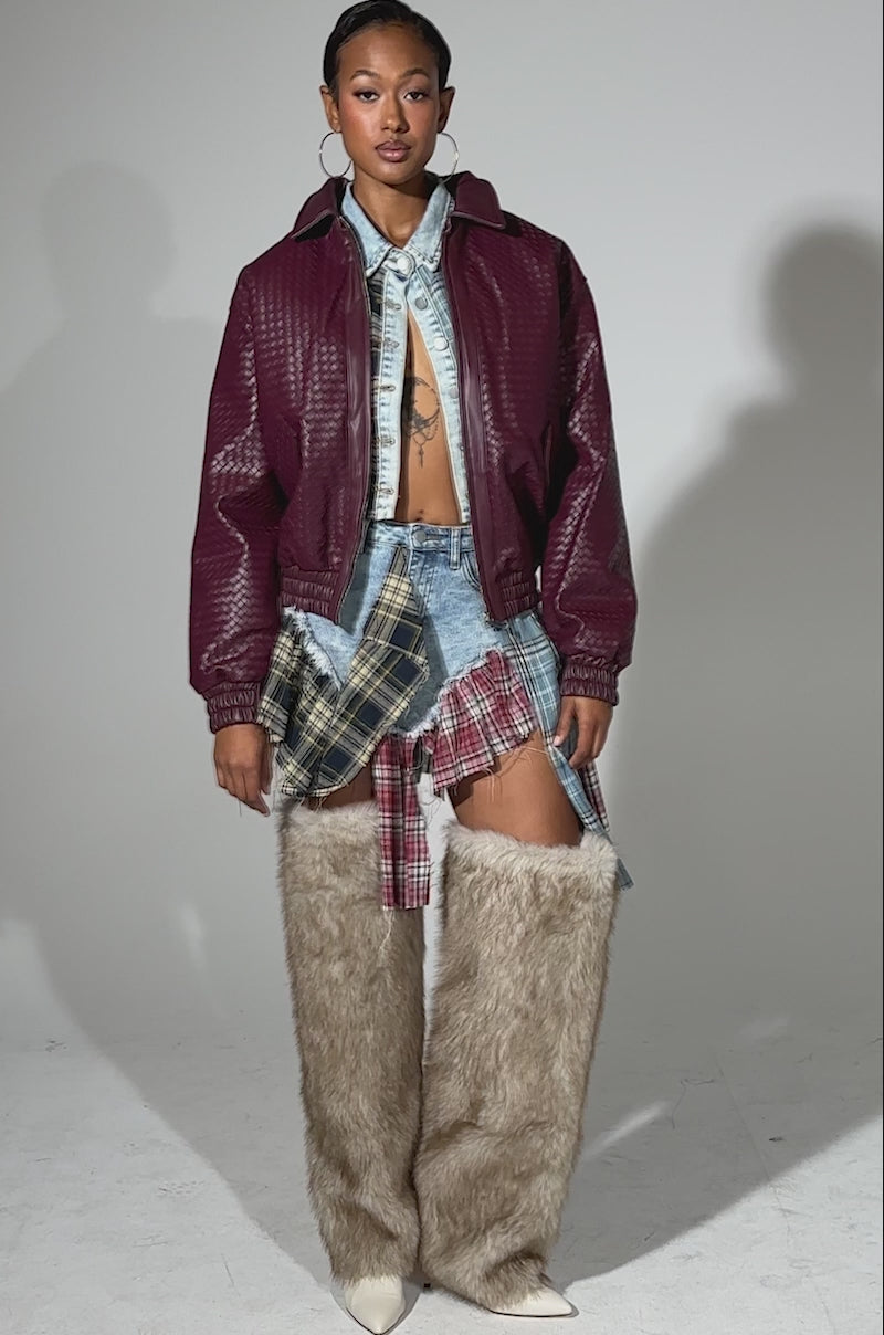 A video of a woman posing confidently in the CHERRY POP BOMBER over a denim shirt and skirt with plaid patches, paired with tall furry beige boots. She accessorizes with hoop earrings and wears her hair styled back.