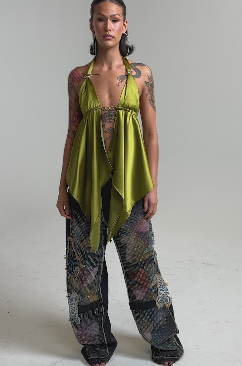 A video of a woman in the BUTTERFLY EFFECT RELAXED DENIM with colorful patchwork and appliqués, paired with a halter-neck olive green top, open-toe shoes, and hoop earrings.