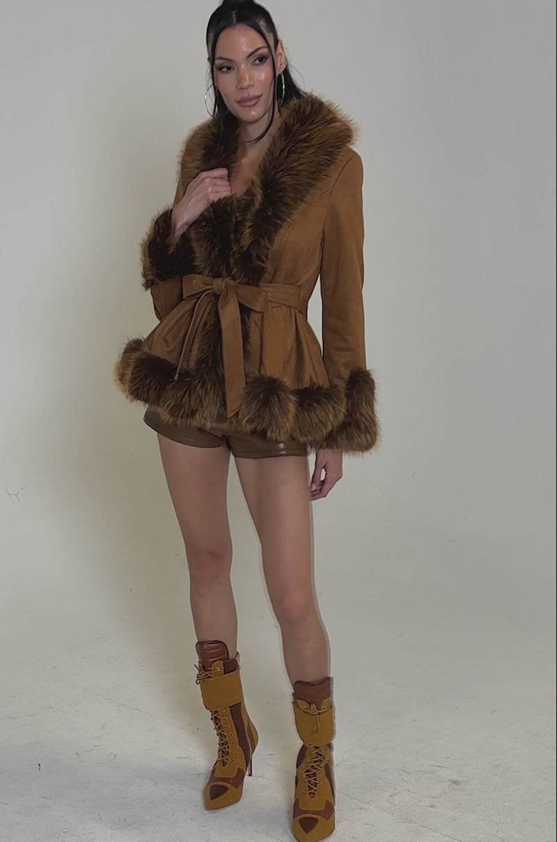A video of a woman posing confidently against a white background wearing the SUEDE KENZY FUR TRIM MOTO jacket, paired with matching brown shorts and tan lace-up chunky heel boots.
