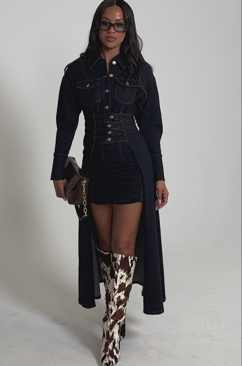 Video of woman modeling the CITY NIGHTS DENIM BUTTON DOWN, a longline dark-wash blouse with silver buttons, paired with a matching skirt, tall cow-print boots, sunglasses, and a small brown shoulder bag.