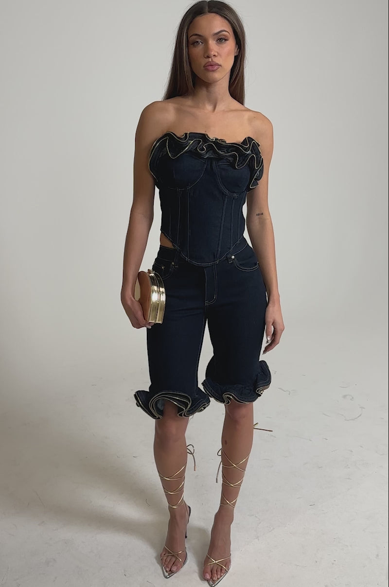 Video of woman modeling the SOMETHING DIFFERENT DENIM CORSET WITH ZIPPER DETAIL and matching high-waisted shorts, holding a clutch purse against a plain light gray background.