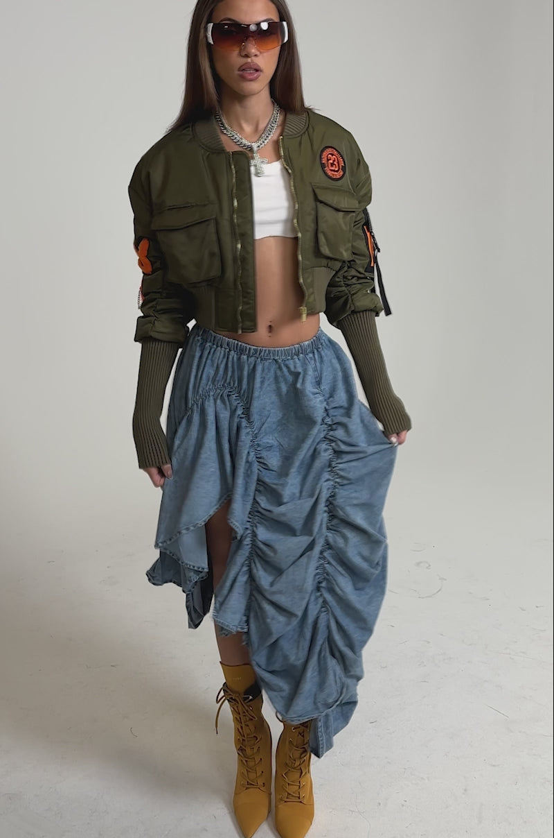 Video of woman modeling the LETS CHANGE IT UP RUCHED MIDI SKIRT IN CHAMBRAY, mustard lace-up high-heeled boots, sunglasses, and layered necklaces.