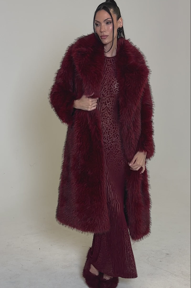 A video of a woman with straight dark hair pulled back stands confidently against a plain background, wearing the OSLO FAUX FUR COAT IN RED MULTI over a matching red dress and red fur shoes.