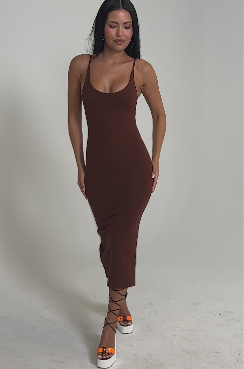 Video of woman modeling the REYNA MIDI BODYCON DRESS IN BROWN, a fitted midi dress with a scoop neckline. She accessorizes with strappy heels and stands against a plain light background, smiling confidently.