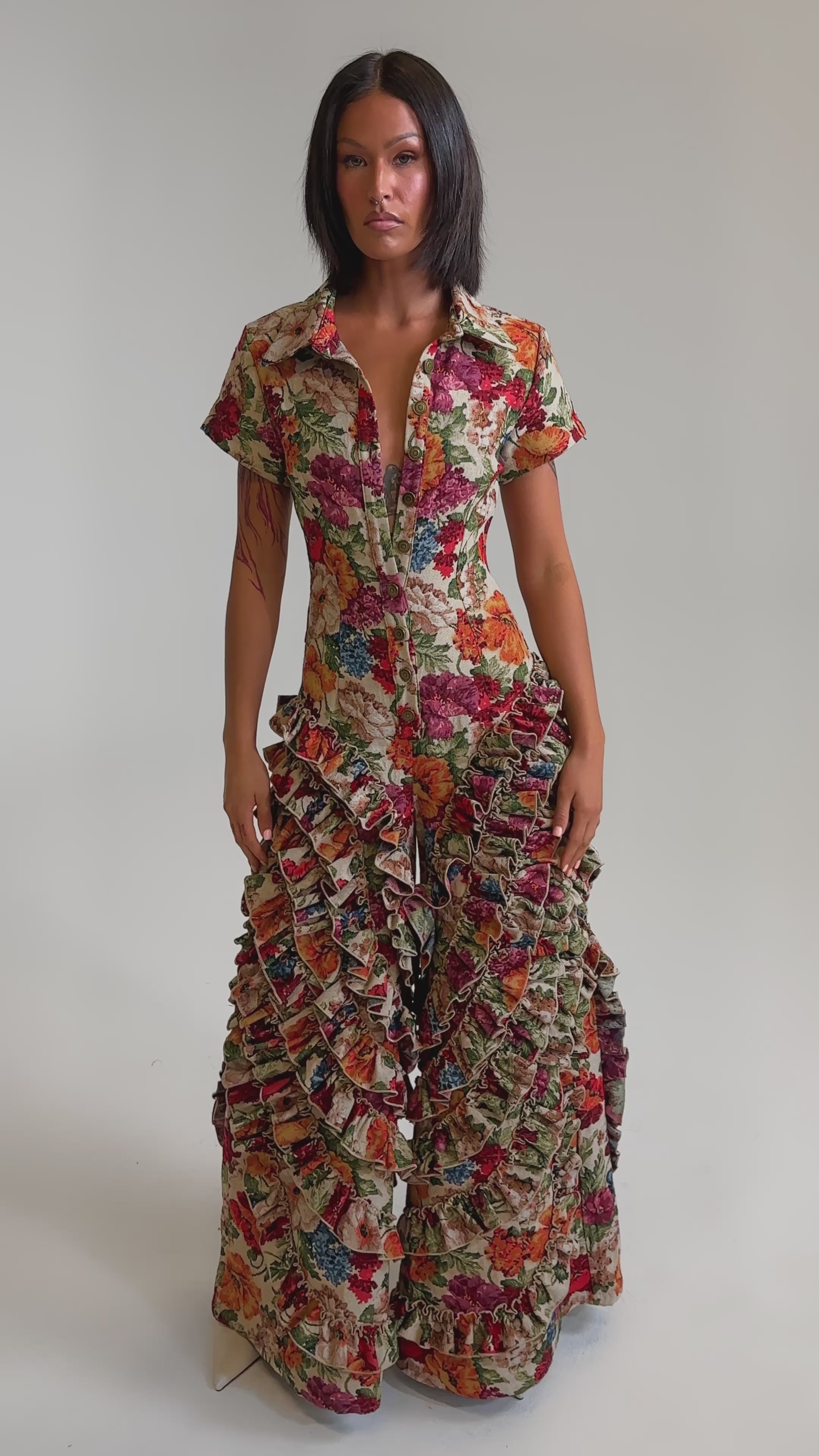 A video of a woman in the CRAZY IN LOVE FLORAL JUMPSUIT, featuring a vibrant, multicolored floral pattern, dramatic ruffle trim from the waist down, structured jacquard fabric, short sleeves, and a deep V-neckline.
