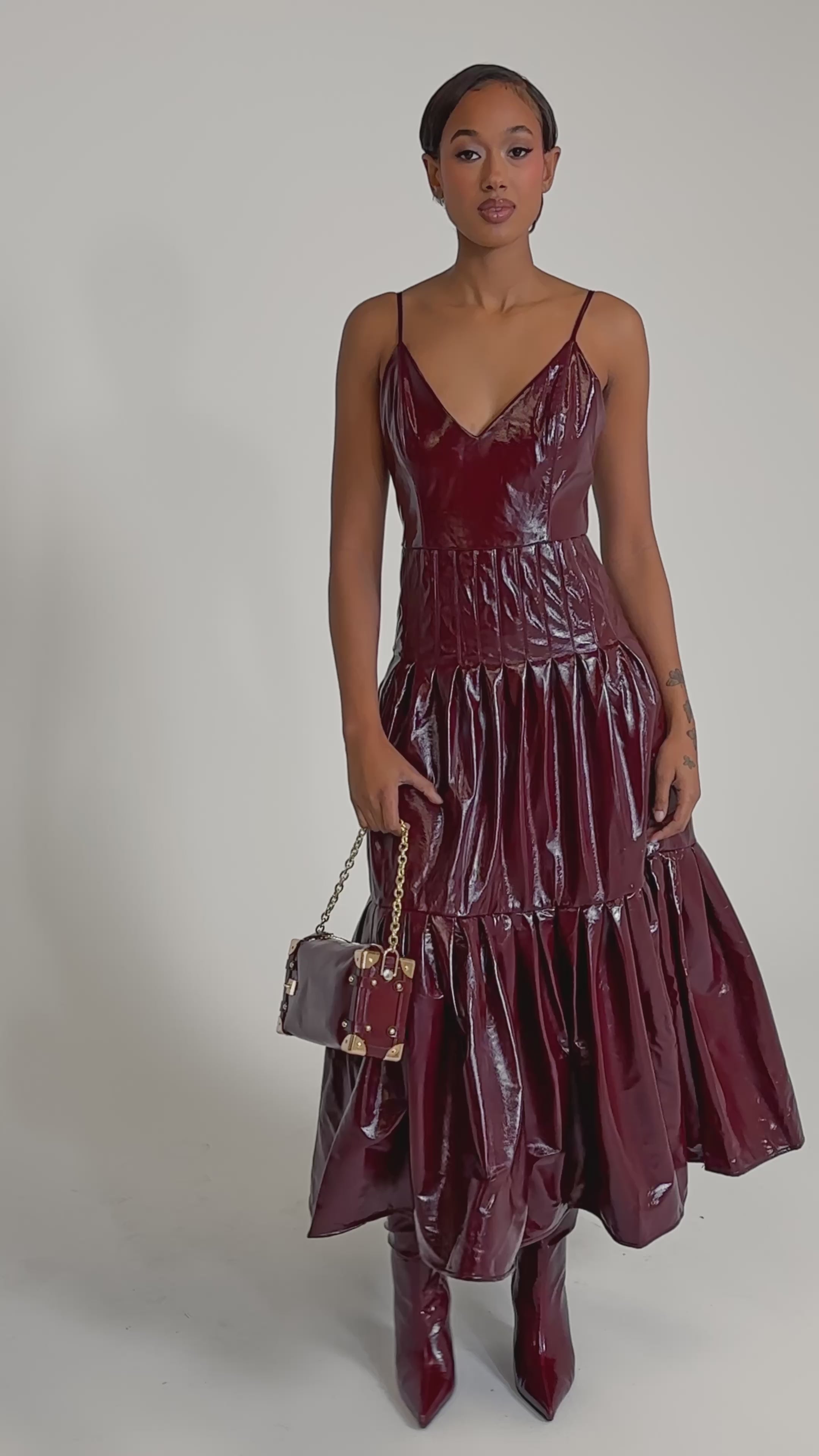 A video of a woman wearing the RACHEL PU MAXI DRESS in shiny burgundy faux leather with a flared skirt, paired with matching knee-high boots and a small handbag.