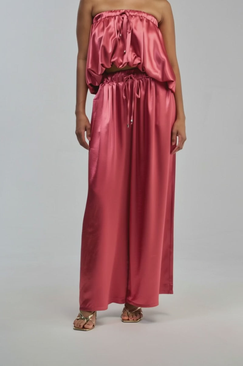 Video of a woman modeling the DREAM GIRL SATIN PANT—a shiny pink, wide-leg pant with a high-rise elastic waistband and a loose fit.