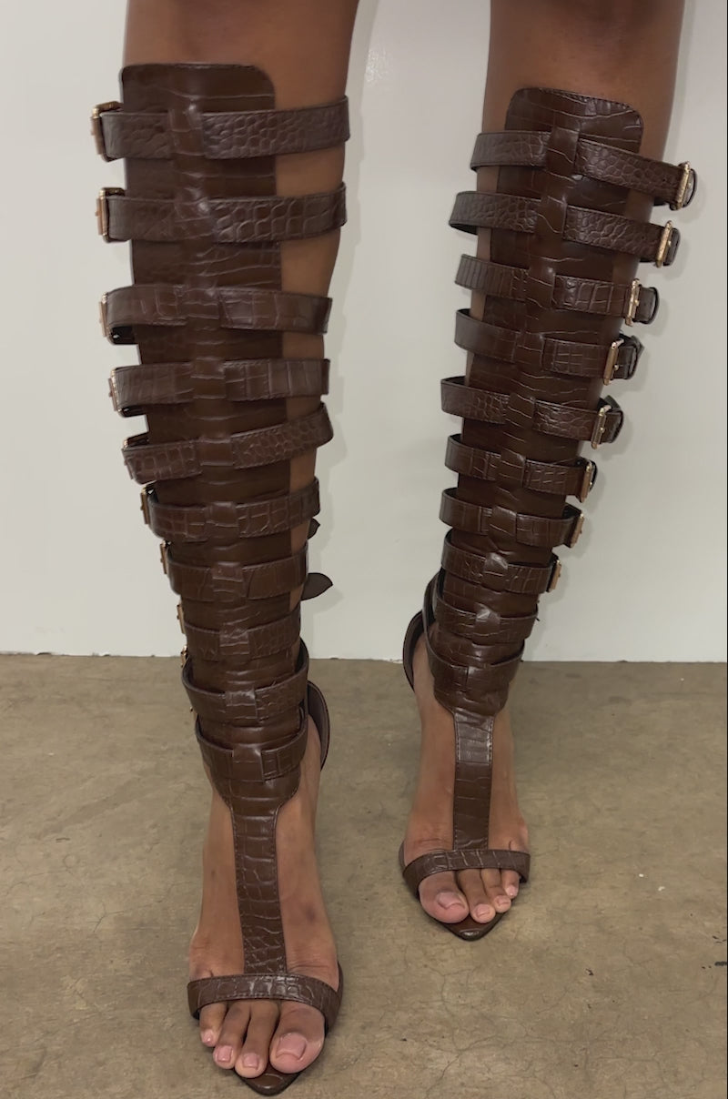 A video of a person wearing a dark brown faux croc-embossed leather mini skirt and AZALEA WANG CHERELLI BROWN CROC OVER THE KNEE SANDAL, posing on a concrete floor against a white background.