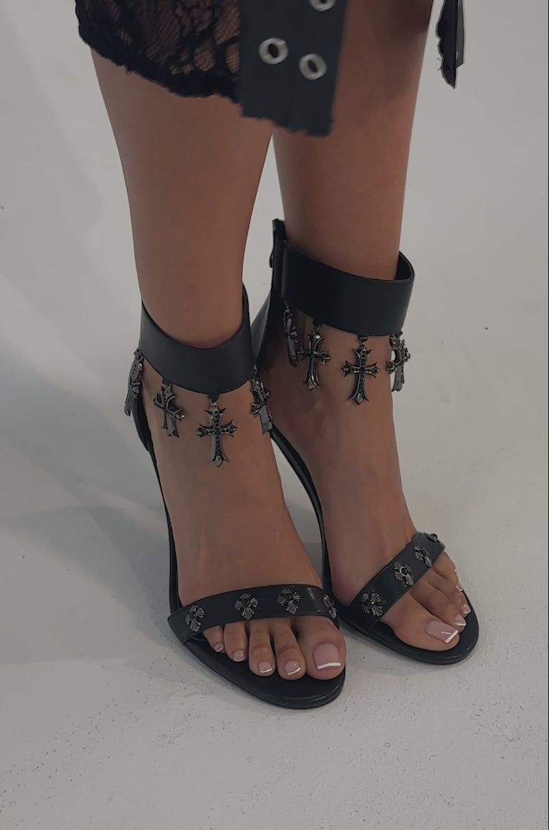 A video of a model wearing the AKIRA GOLD CLERGY BLACK EMBELLISHED SEXY STRAP STILETTO HEEL, featuring ankle straps with metallic cross charms, decorative studs, and open toes against a simple light background.