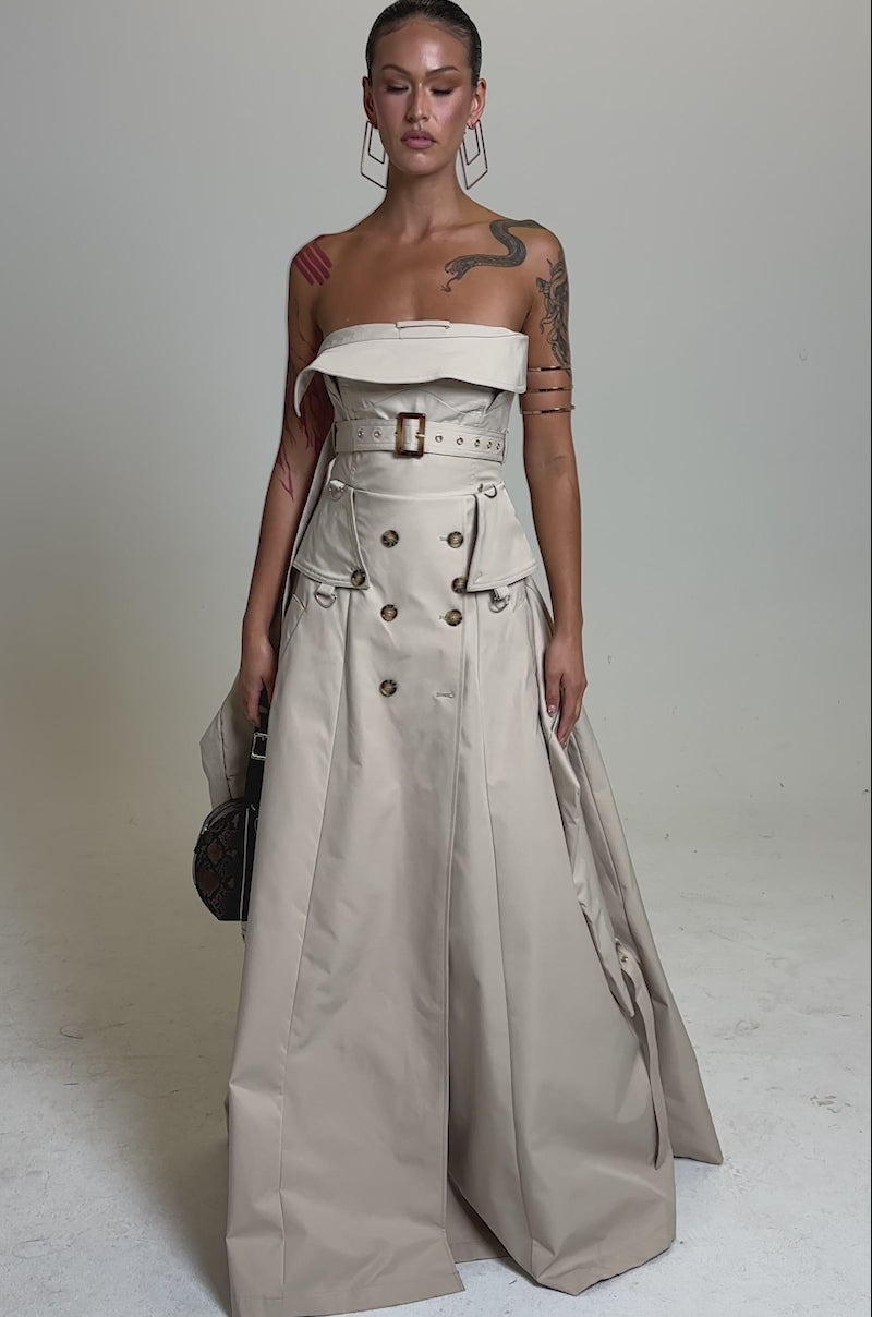 A video of a woman wearing the STRAPLESS TRENCH MAXI DRESS in beige, featuring button and belt details, styled like a trench coat. She carries a round, brown patterned handbag and displays arm tattoos, set against a plain light gray background.