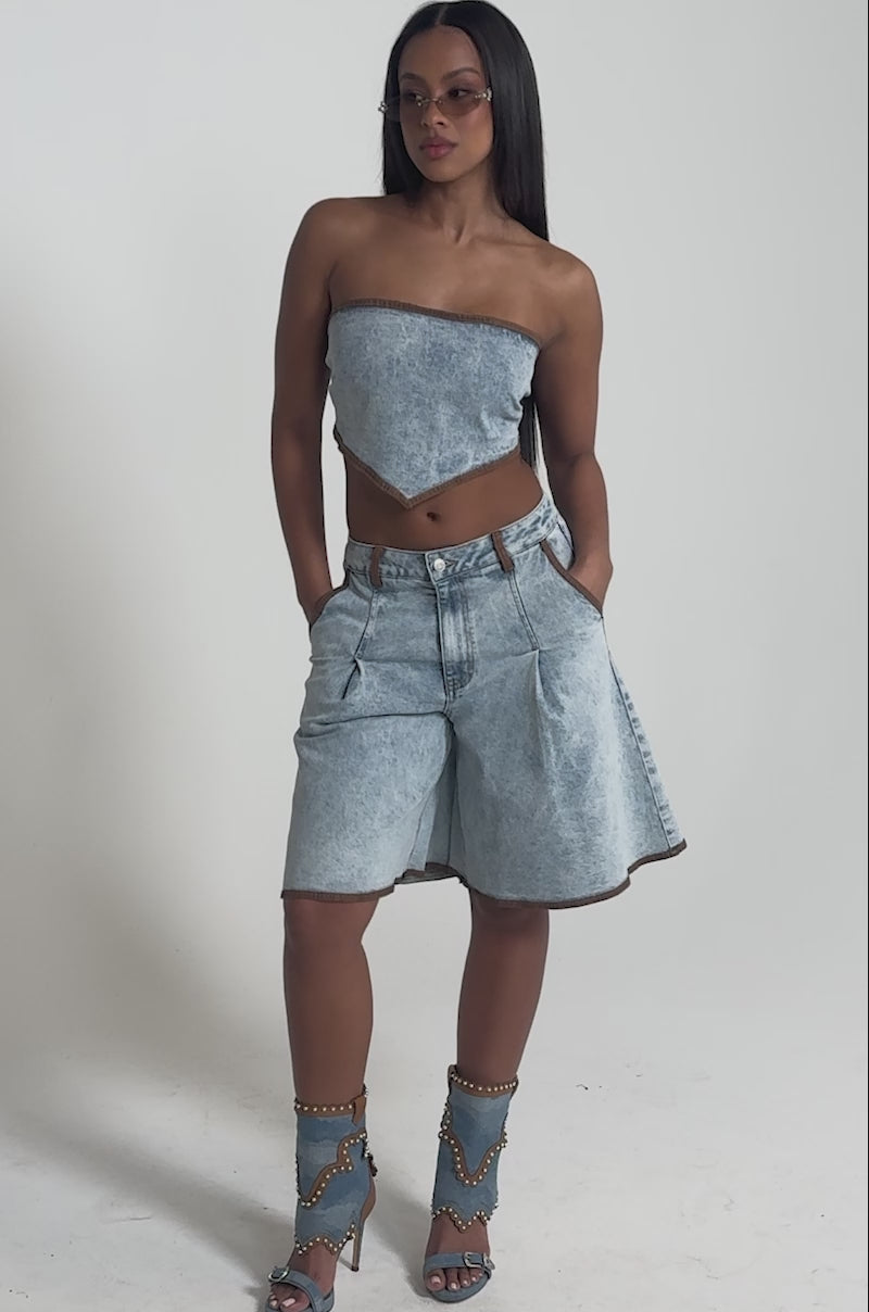 A video of a woman wearing the TOO COOL FOR THIS CORSET IN DENIM, pairing it with wide-leg denim shorts and brown-trimmed studded festival boots. 