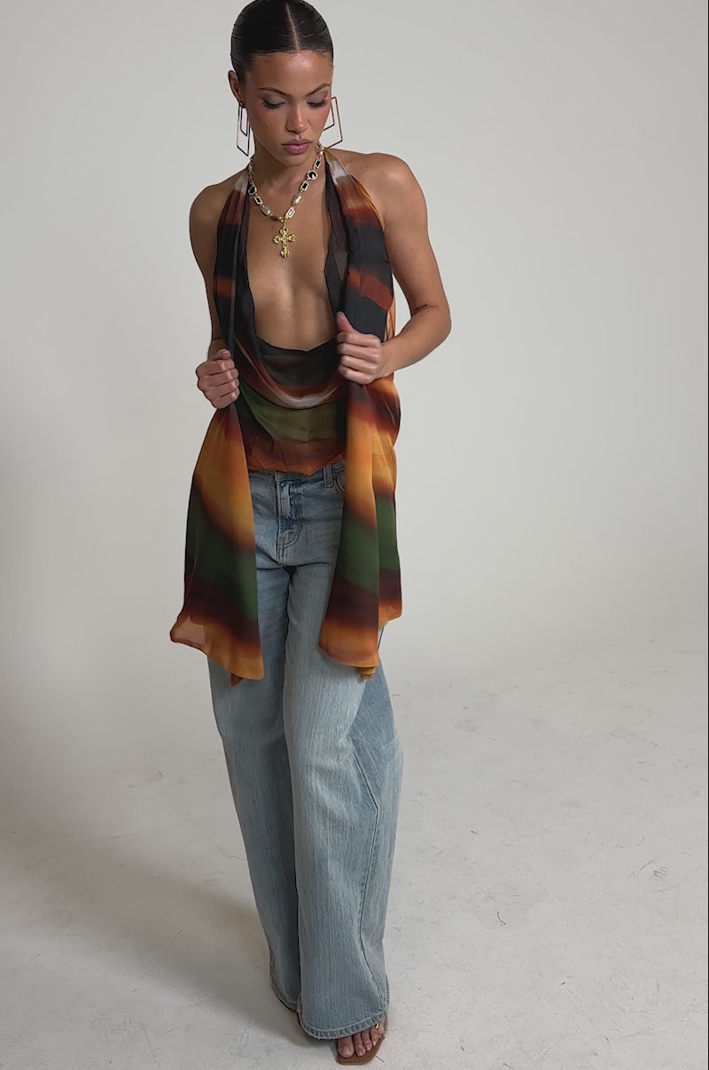 Video of woman modeling the SELENA DEEP COWL HALTER TOP, a colorful striped halter with a deep cowl neckline. She pairs it with light blue jeans and stands against a neutral background