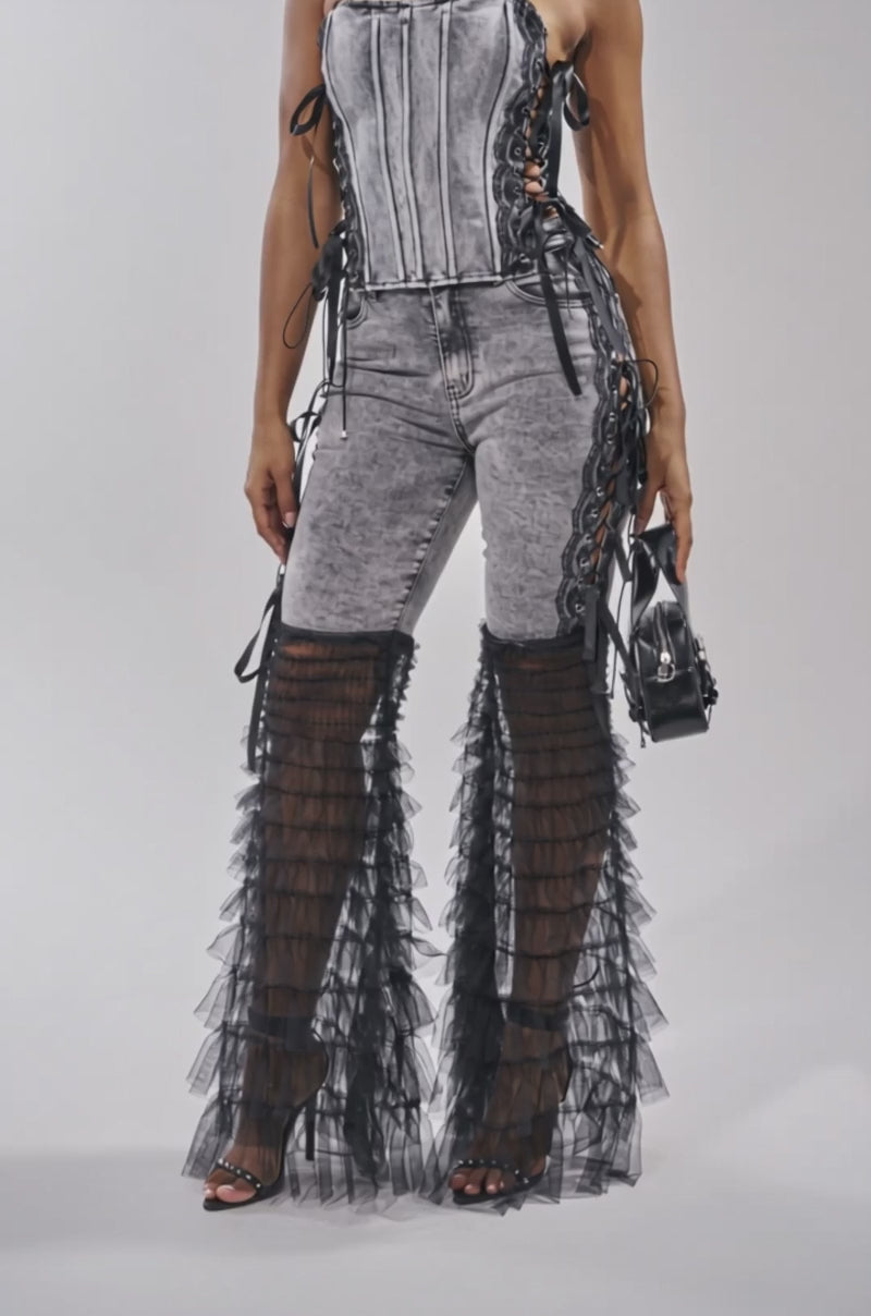A video of a woman in the TICKED AND TIED LACE DETAIL DENIM CORSET, styled with fitted grey jeans featuring bold black sheer ruffled flares, a small black handbag, and black heels.