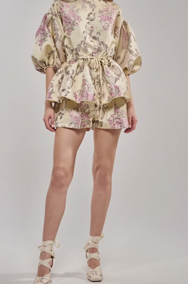 Video of woman modeling the PORCELAIN DOLL BROCADE TOP, a cream-colored floral print puff-sleeve blouse with a matching short and belted waist, posing against a plain light background.