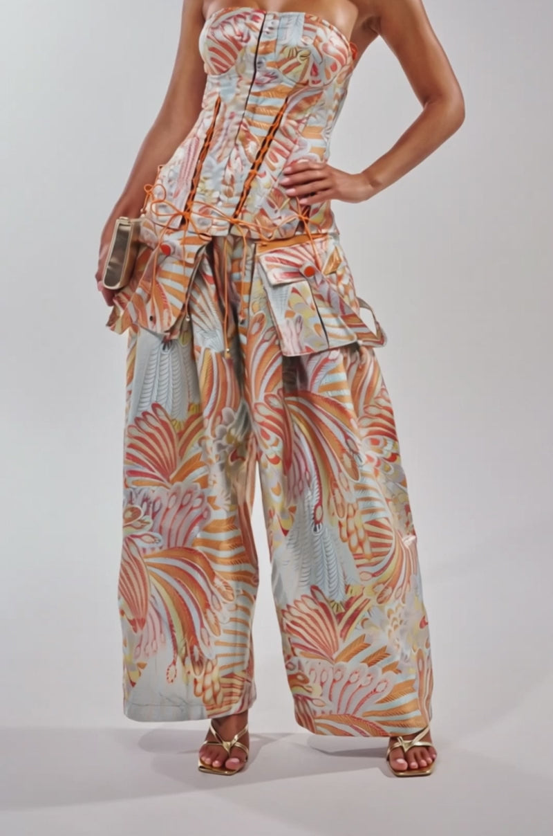 A video of a woman wearing the MARTHA FLORAL TROUSER—blue wide-leg pants with a high-rise waistband and subtle metallic accents—paired with a strapless corset top, clutch, and strappy heels. 