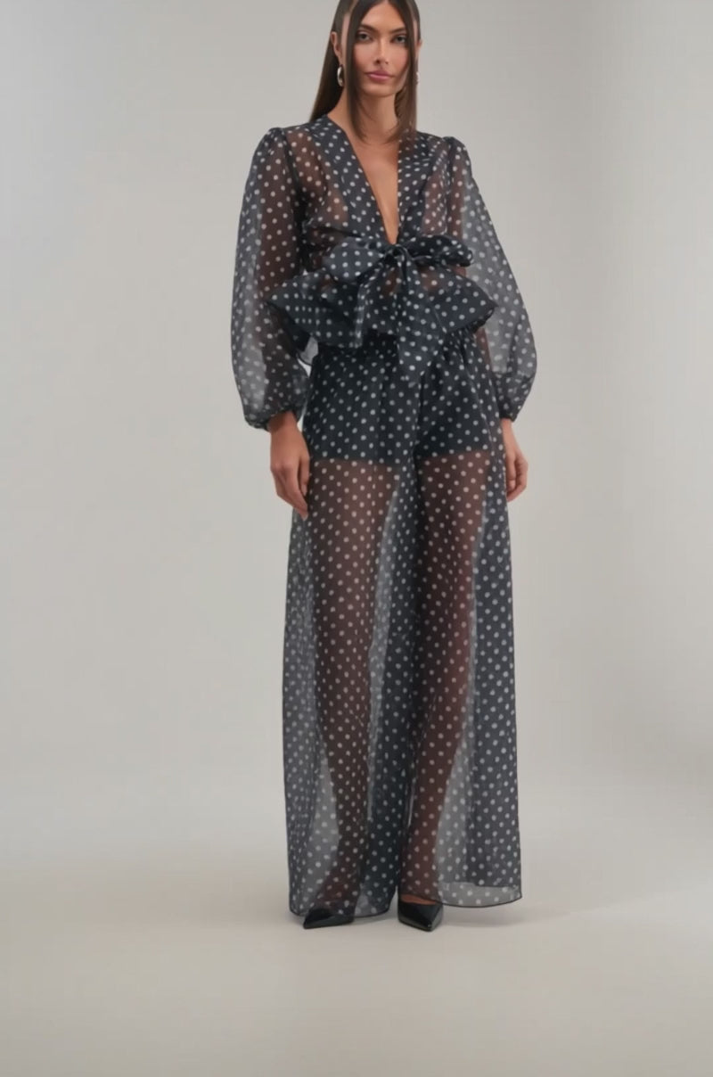 A video of a woman wearing the PRETTY GIRL POLKA DOT PALAZZO PANT, featuring sheer black polka dots, a deep V-neck, puffed long sleeves, a large waist bow, black undergarments, and flowing palazzo pants beneath a floor-length overlay.