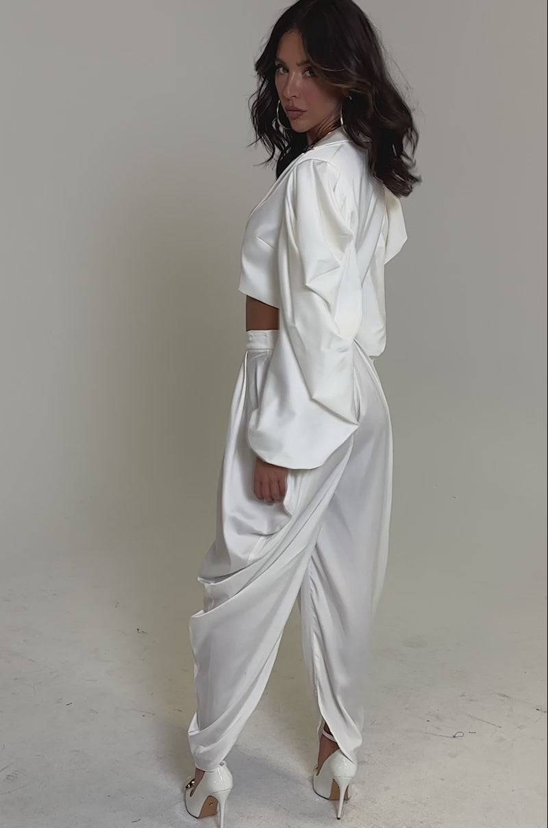 Video of woman modeling the NEW BEGINNING CROPPED BLAZER in white, featuring billowy sleeves and paired with wide-leg pants. She accessorizes with a chunky gold necklace, hoop earrings, and holds a small gold clutch against a plain background.