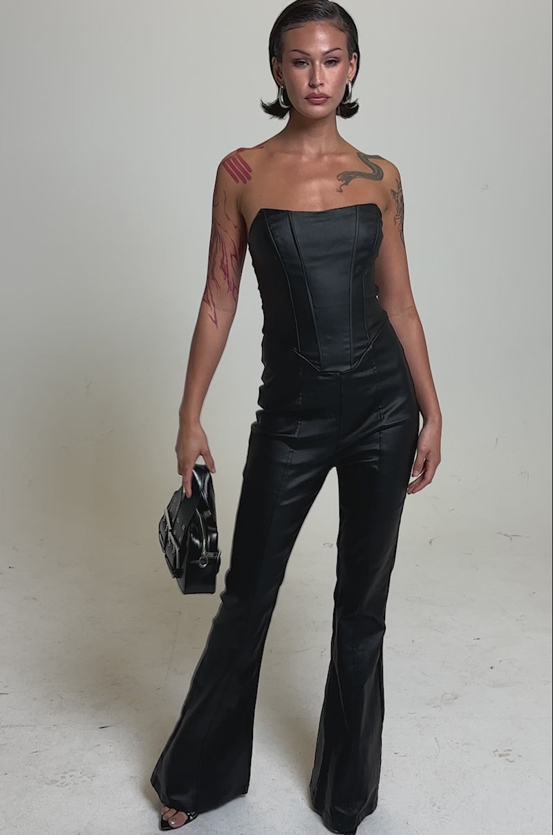 A video of a person with short dark hair modelling the HOT GIRL PU JUMPSUIT, paired with a small black handbag. Tattoos accent their arm and shoulder as they stand confidently before a plain light background.