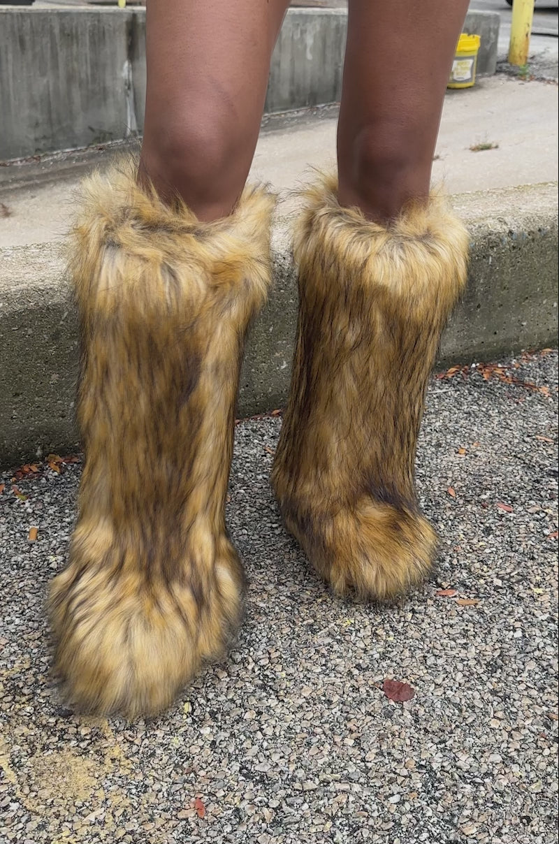 A video of a person wearing the AZALEA WANG FAYES BROWN MULTI FURRY KNEE HIGH FLAT BOOTS standing outdoors on a concrete surface near a yellow railing.