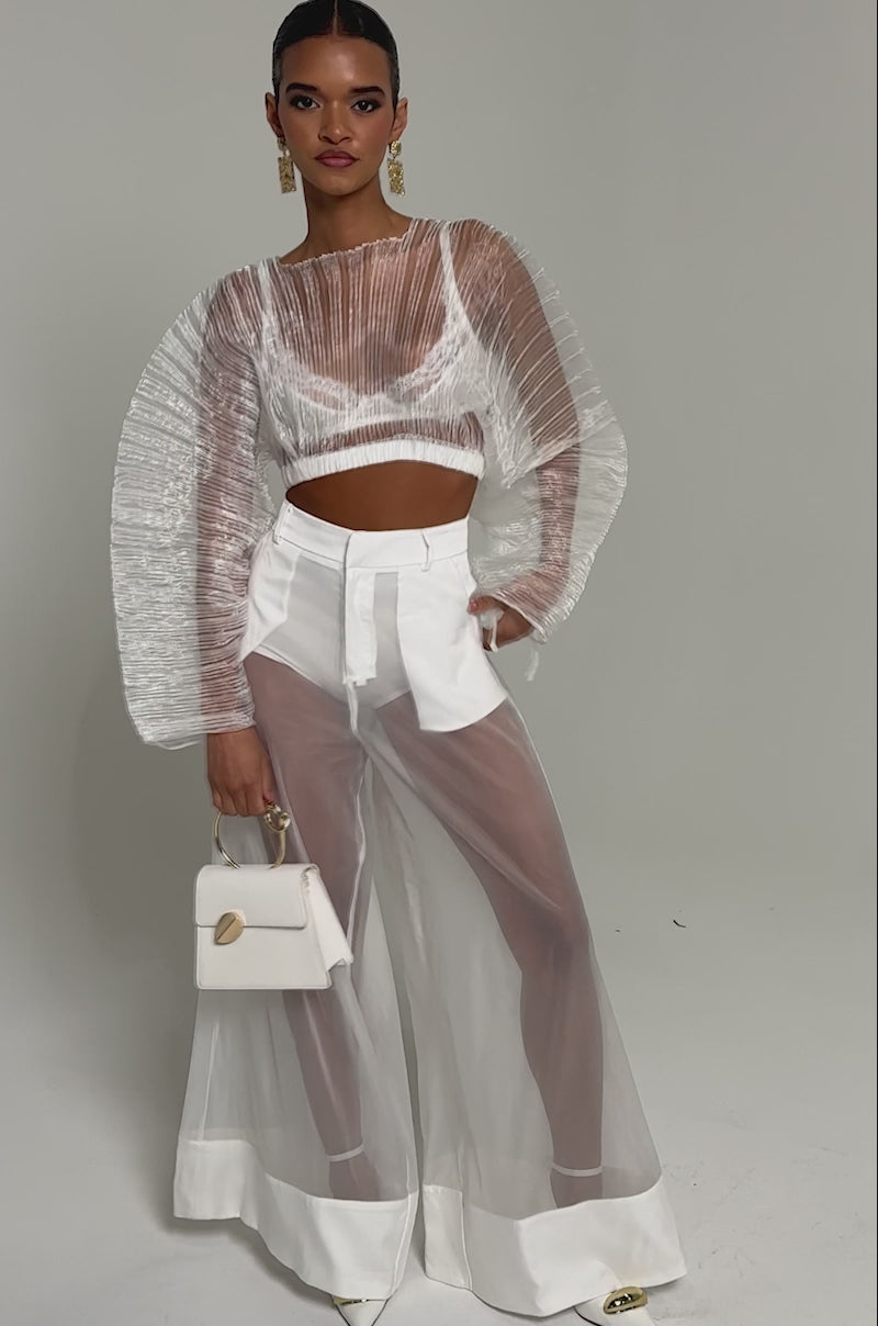 Video of woman modeling the VACATION MODE PLEATED BLOUSE—sheer and dramatic with curved sleeves—over a white bralette and high-waisted trousers, styled with gold earrings, a small bag, and slicked-back hair against a gray backdrop.