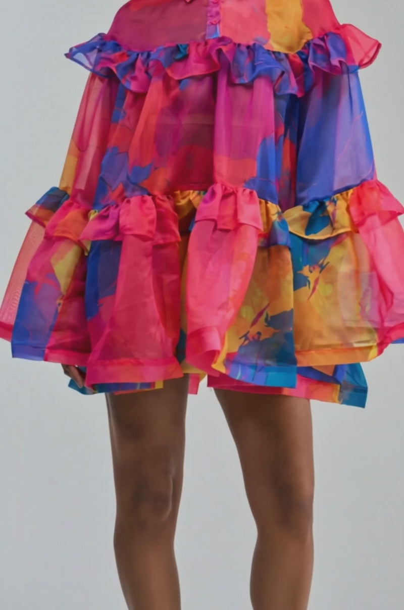 Video of a woman modeling the CALL ME BABY CHIFFON MINI DRESS IN PINK MULTI—a sheer, colorful ruffle mini dress featuring bold pink, blue, and orange abstract patterns and a buttoned collar.