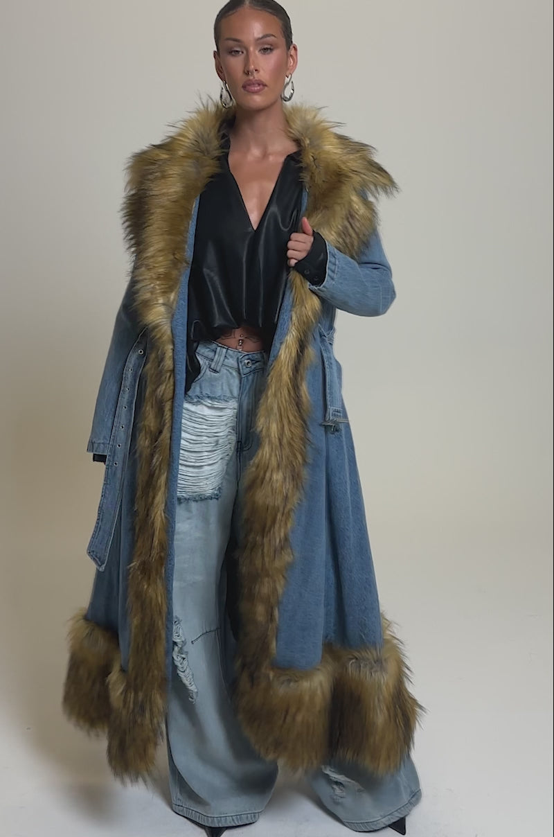Video of woman modeling the VIVIAN FUR TRIM TRENCH IN DENIM, a blue denim coat with tan faux fur trim on the collar, front, and hem. She pairs it with wide-leg denim pants against a plain light gray background, her hair pulled back.