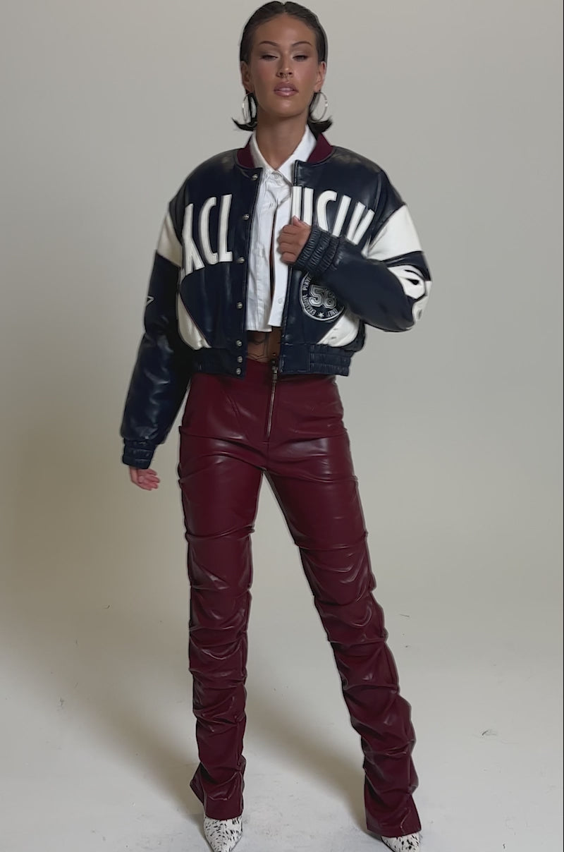 A video of a woman in the Down and Dirty Faux Leather Pant—high-waisted, burgundy, with a skinny-leg fit and ruched details—paired with a two-tone varsity jacket, cropped white top, and white pointed shoes against a plain backdrop.