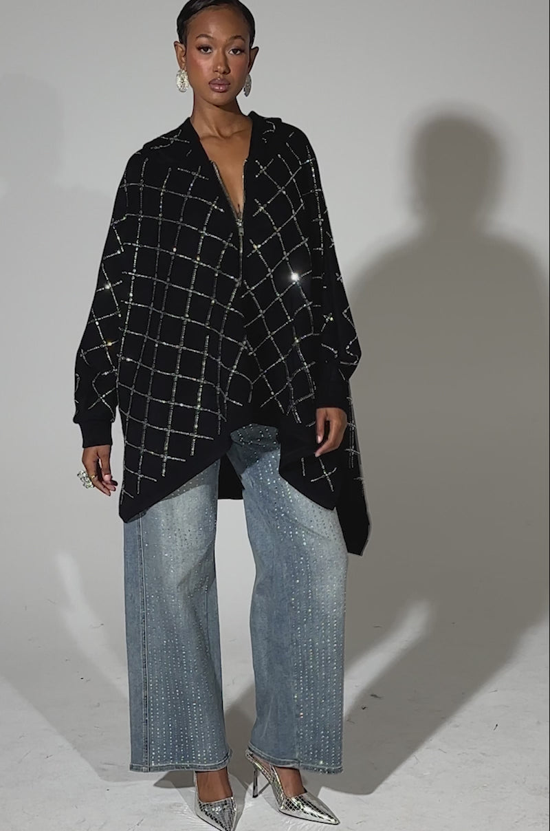 A video of a woman in streetwear, wearing the BLING BLING OVERSIZED ZIP UP SWEATSHIRT, wide-leg blue jeans with sparkling embellishments, silver pointed-toe heels, and statement rings.