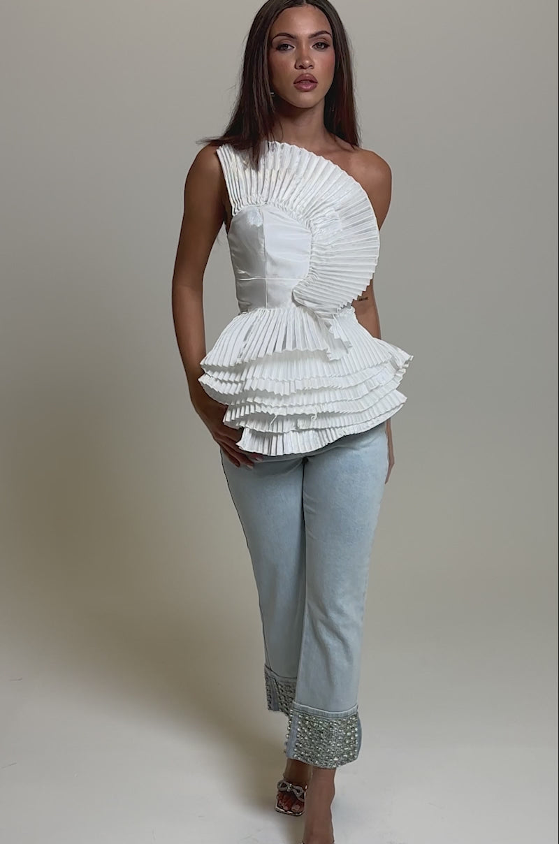 Video of woman modeling the SUNSHINE PLEATED RUFFLE BLOUSE—a chic white top with layered pleats—paired with light blue jeans, standing against a plain light gray background.