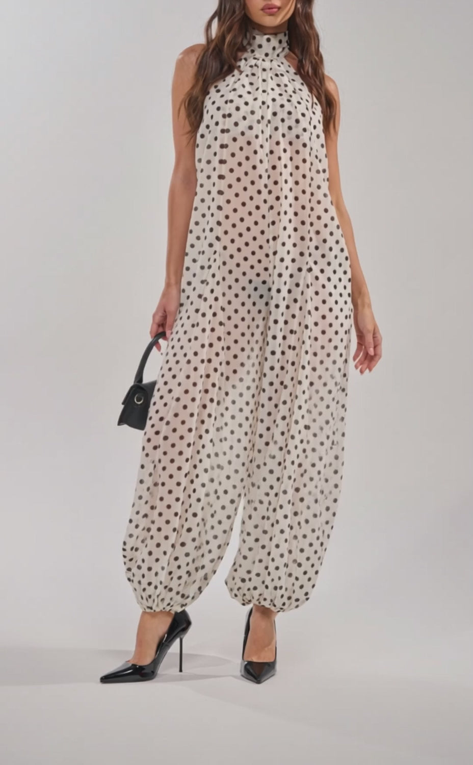 A video of a woman in glasses wearing the GIRLY POP POLKA DOT JUMPSUIT with a small black handbag and black pointed heels.